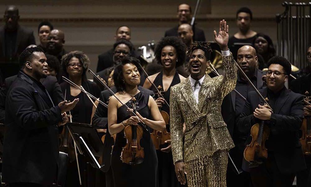 站着的非洲裔管弦乐队音乐家, 在癌症舞台上鼓掌，身穿金色服装的钢琴家挥手致意.