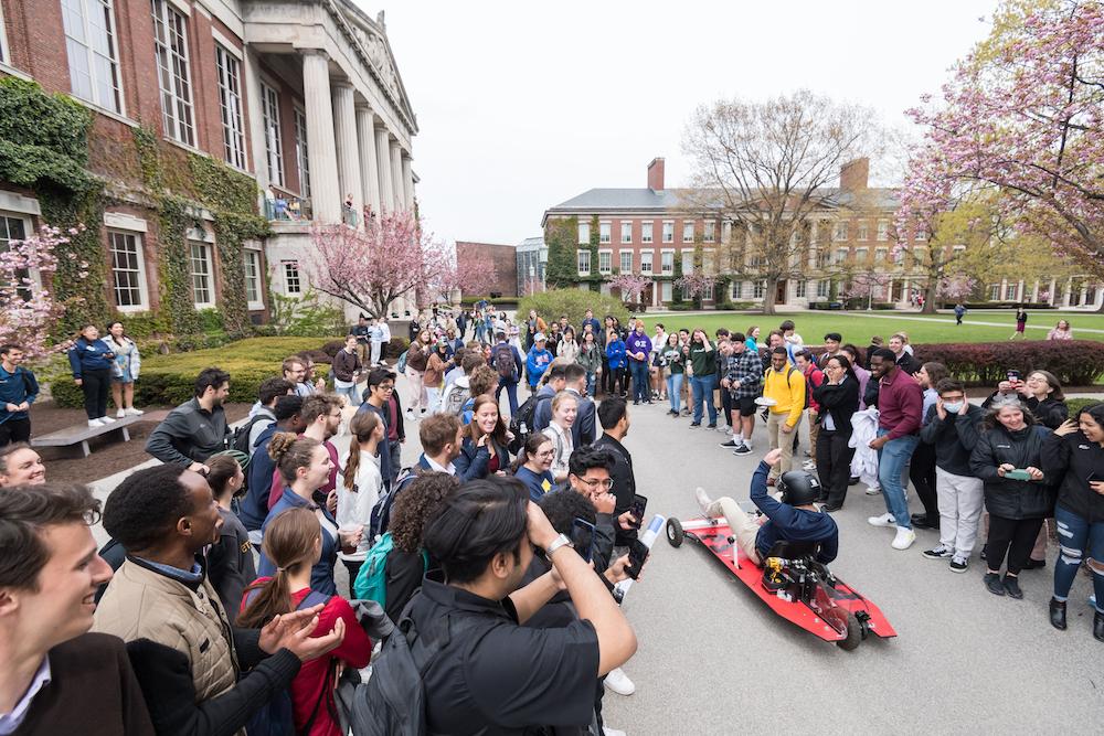 College student in a car powered by a hand drill is cheered on while he races by a group of supporters
