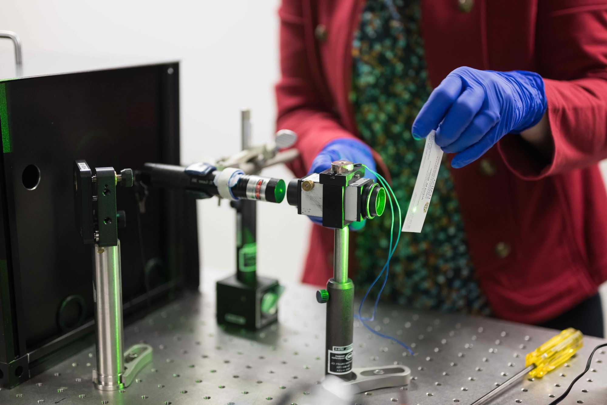 A scientist's gloved hands holds a material in front of a set-up of pulsed lasers to create catalysts.
